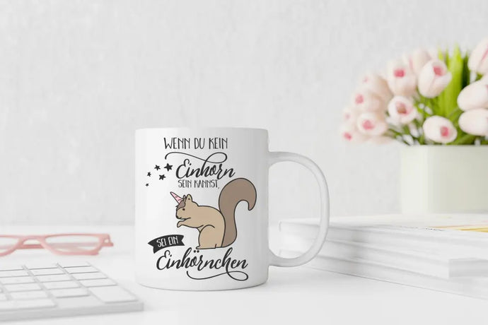 Ein humorvolles Bild einer Tasse mit dem Schriftzug 'Wenn du kein Einhorn sein kannst, sei ein Einhörnchen' und einem niedlichen Einhörnchen-Motiv. Die Tasse steht auf einem Tisch und strahlt Spaß und Einzigartigkeit aus.